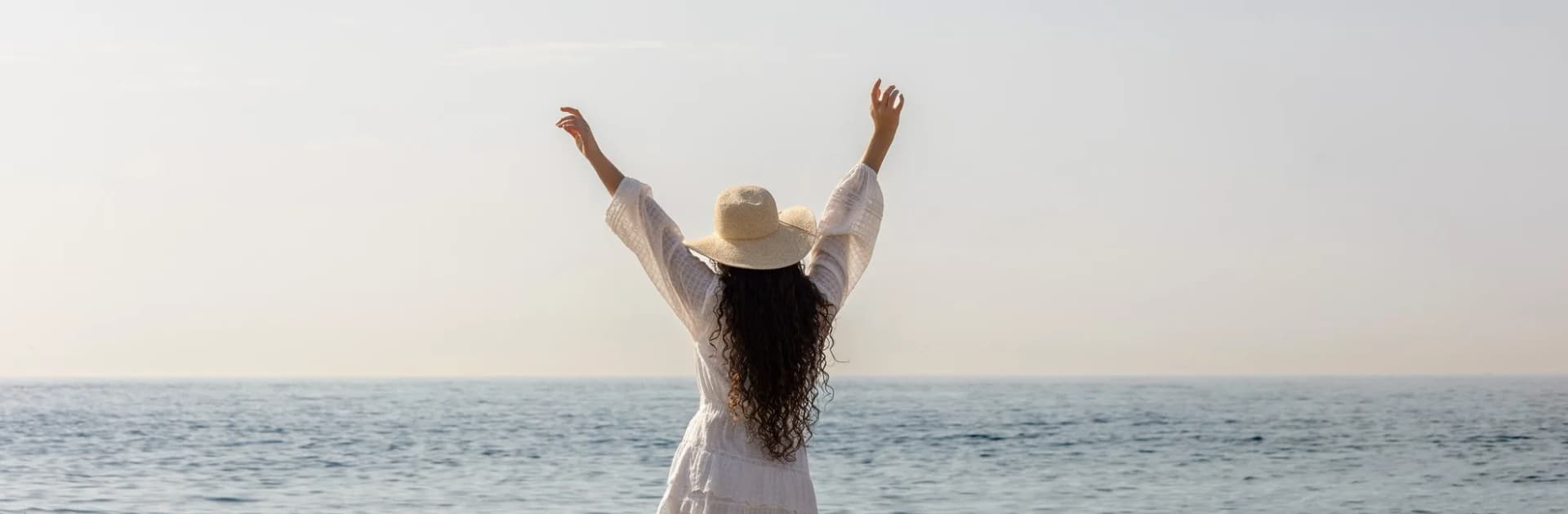 Woman in a sun hat, arms raised, facing the sea